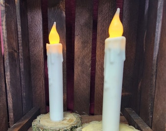 May include: Two white pillar candles with a warm yellow glow, sitting on rustic wooden slices with bark.