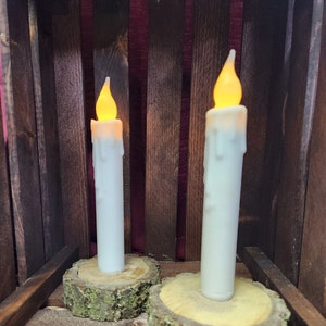 May include: Two white pillar candles with a warm yellow glow, sitting on rustic wooden slices with bark.