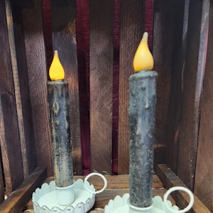 May include: Two black taper candles with white candle holders. The candle holders have a scalloped edge and a handle. The candles are sitting on a wooden surface.