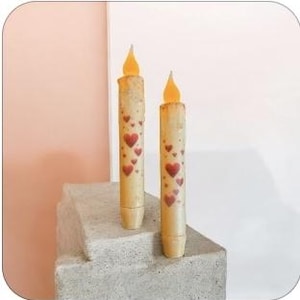 May include: Two battery-operated, cream-colored candles with a heart pattern. The candles have a flickering, flame-shaped LED at the top. The candles are approximately 8 inches tall and are displayed on a concrete block.