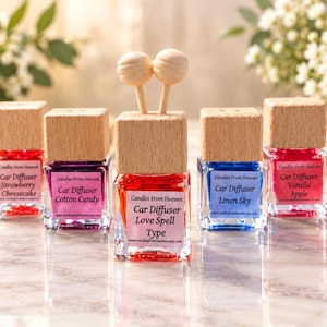 May include: Five square glass car diffuser bottles with light wood caps and wooden sticks. The labels read Candles From Heaven, Car Diffuser, and the scents Strawberry Cheesecake, Cotton Candy, Love Spell Type, Linen Sky, and Vanilla Apple.