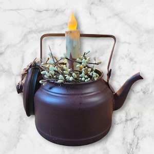 May include: A decorative, burgundy-colored teapot with a handle and spout. Inside the teapot is a battery-operated candle with a star design, and sprigs of white berries. The teapot's lid is open, revealing the interior.