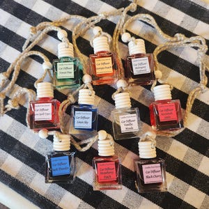 May include: A collection of ten small glass bottles with string hangers. Each bottle contains a different scented oil and has a label that reads "Car Diffuser" followed by the scent name. The scents include Forest Pine, Georgia Peach, Mahogany Teakwood, Love Spell Type, Linen Sky, Vanilla Woods, Macintosh Apple, Dark Thunder, Strawberry Patch, and Black Cherry.