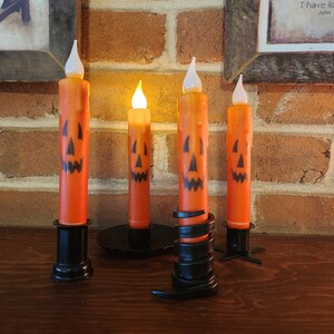 May include: Four orange Halloween-themed LED candles with black jack-o'-lantern faces. The candles are in black metal holders, with one candle lit. The background includes a brick wall and framed artwork.