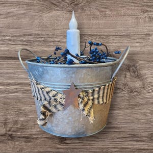 May include: A galvanized metal bucket with a decorative star and ribbon. The bucket holds a white LED candle and blue berry stems. The bucket has two metal handles and is set against a wood grain background.