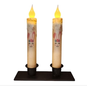 May include: Two white pillar candles with bunny faces and floral headbands on them. The candles are lit and are on a black metal candle holder.
