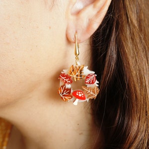 May include: A pair of gold dangle earrings with a white circular base and red and orange leaves and mushrooms.