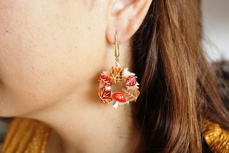 May include: A pair of gold dangle earrings with a white circular base and red and orange leaves and mushrooms.