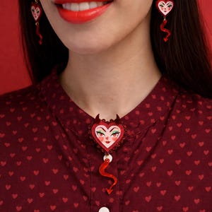 Peut inclure: Un ensemble de bijoux rouges en forme de cœur avec des visages diaboliques et des accents rouges. Les boucles d'oreilles pendantes ont une queue rouge incurvée. La broche est épinglée sur une chemise bordeaux à motifs de cœurs.