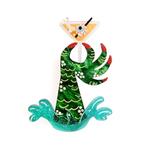 May include: A green monster hand with red claws holding a martini glass with an eyeball and a straw. The monster hand is emerging from blue water.