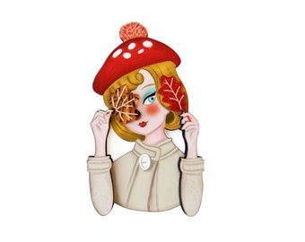 May include: A wooden brooch depicting a cartoon woman with blonde hair wearing a red mushroom hat and holding two autumn leaves in front of her face.