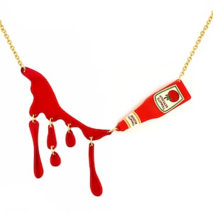 Op de afbeelding: Een goudkleurige ketting met een rode acryl hanger in de vorm van een gekantelde ketchupfles met het label Tomato Ketchup en Tomato Blood, verbonden met een rood druipend vloeistofontwerp met vier bungelende roodkleurige traanvormige bedels.
