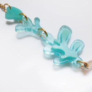 Puede incluir: Un collar de color azul turquesa con un acabado brillante. El collar presenta una serie de formas abstractas, incluyendo un coraz&oacute;n y un dise&ntilde;o en forma de hoja. El collar est&aacute; hecho de acr&iacute;lico y tiene una cadena dorada.