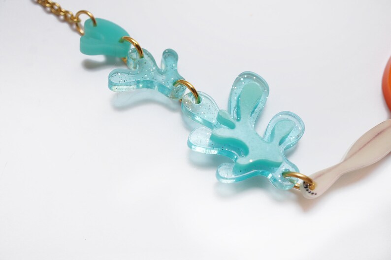 Puede incluir: Un collar de color azul turquesa con un acabado brillante. El collar presenta una serie de formas abstractas, incluyendo un coraz&oacute;n y un dise&ntilde;o en forma de hoja. El collar est&aacute; hecho de acr&iacute;lico y tiene una cadena dorada.