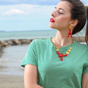 May include: A woman wearing a green and white striped t-shirt with a red lobster necklace and matching earrings. The necklace is made of gold and red lobster charms.
