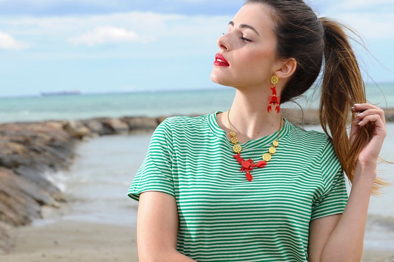 May include: A woman wearing a green and white striped t-shirt with a red lobster necklace and matching earrings. The necklace is made of gold and red lobster charms.