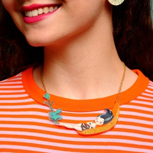 Puede incluir: Un collar de cadena dorada con un colgante colorido que representa a una mujer de dibujos animados tumbada en una luna creciente. El colgante est&aacute; hecho de pl&aacute;stico y tiene un acabado brillante.