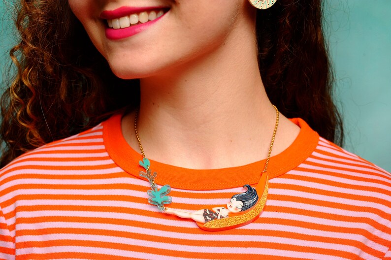 Puede incluir: Un collar de cadena dorada con un colgante colorido que representa a una mujer de dibujos animados tumbada en una luna creciente. El colgante est&aacute; hecho de pl&aacute;stico y tiene un acabado brillante.