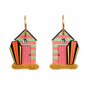 May include: A pair of pink and green beach hut earrings with gold hoops. Each earring features a surfboard and the number "13" on the front of the hut.