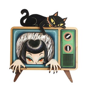 Può includere: Un oggetto da parete in legno che raffigura un televisore in stile vintage con cornice in legno chiaro e pannello di controllo verde. Un gatto nero è posato sopra, mentre un personaggio con capelli neri e occhi rossi emerge dallo schermo.