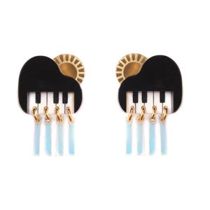 Puede incluir: Un par de pendientes caprichosos con un diseño de piano negro con un teclado blanco y negro, un sol dorado y detalles colgantes en azul claro. Los pendientes están sobre un fondo blanco.