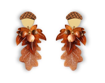 May include: A pair of autumnal earrings. Each earring has a wooden acorn top, a gold-coloured disc, and a cluster of brown and peach-coloured beads. The earrings feature oak leaf-shaped pendants with glittery accents.