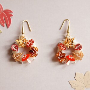 May include: A pair of gold tone earrings with a white circular base and red and gold autumn leaves and mushrooms.
