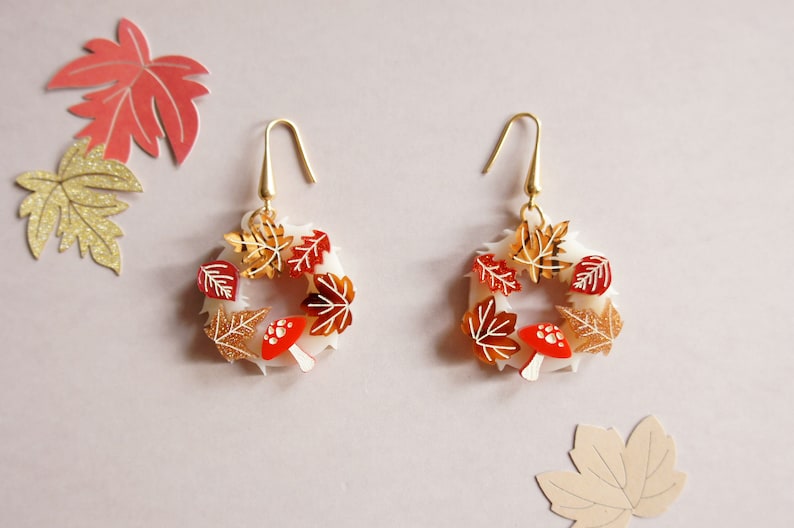 May include: A pair of gold tone earrings with a white circular base and red and gold autumn leaves and mushrooms.