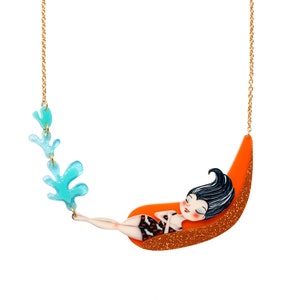 Puede incluir: Un collar de cadena dorada con un dise&ntilde;o de algas azul y verde y un colgante de una mujer durmiendo en una hamaca naranja.