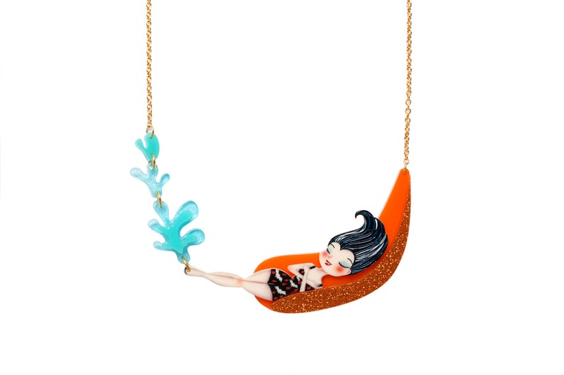 Puede incluir: Un collar de cadena dorada con un dise&ntilde;o de algas azul y verde y un colgante de una mujer durmiendo en una hamaca naranja.