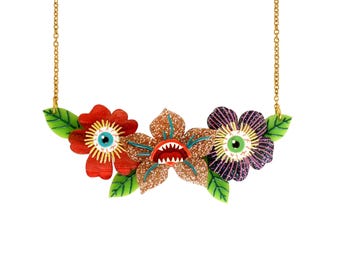 Puede incluir: Un collar de cadena dorada con tres dijes de flores. Las flores son rojas, moradas y doradas con purpurina. Cada flor tiene un ojo grande y una boca.