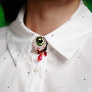 Puede incluir: Una camisa blanca con cuello adornada con pequeños adornos dispersos. Un broche con forma de globo ocular con iris verde, vasos sanguíneos rojos y gemas rojas en forma de lágrima está prendido a la camisa.