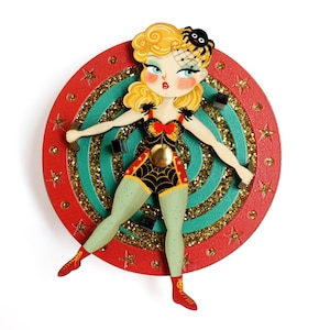 May include: A quirky, vintage-style pin depicting a cartoon woman in a spiderweb costume, with a spider on her blonde hair. She is set against a red and teal target design with gold glitter accents. The pin is a unique, eye-catching accessory.