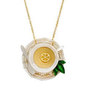 Puede incluir: Un collar de cadena dorada con un colgante de porcelana blanca con forma de taza de té y platillo con una rodaja de limón en el té. El platillo tiene un borde dorado y un diseño floral dorado.