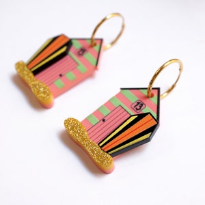 May include: A pair of gold hoop earrings with pink and green beach hut charms. The charms have black stripes, orange and yellow accents, and the number "13" on the front. The bottom of the charms have gold glitter.