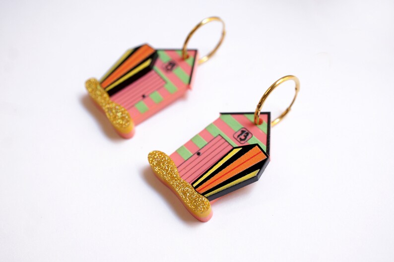 May include: A pair of gold hoop earrings with pink and green beach hut charms. The charms have black stripes, orange and yellow accents, and the number "13" on the front. The bottom of the charms have gold glitter.