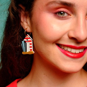 May include: A pair of gold hoop earrings with a red, white, and blue beach hut charm. The charm has a number "13" on the side.