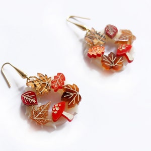 May include: A pair of gold hoop earrings with a fall-themed design. The earrings are made of clear plastic and feature red and orange leaves, mushrooms, and glitter accents.