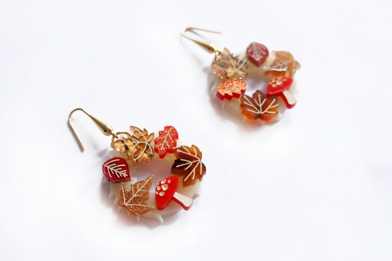 May include: A pair of gold hoop earrings with a fall-themed design. The earrings are made of clear plastic and feature red and orange leaves, mushrooms, and glitter accents.