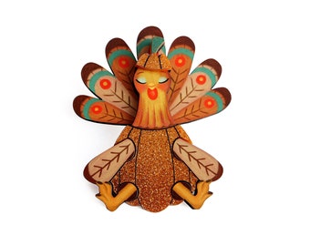 May include: A wooden turkey ornament with a glittery brown body, orange beak, and teal and brown feathers. The turkey has closed eyes and a small brown hat on its head.