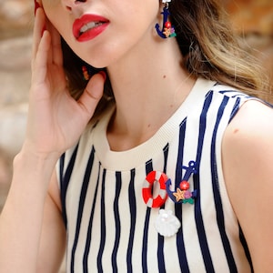 以下が含まれることがあります： 白と青のストライプのシャツを着た女性が、ライフセーバー、アンカー、貝殻をモチーフにしたマリンテイストのブローチをつけています。彼女は、貝殻のチャームが付いた青と白のアンカーのイヤリングをしています。