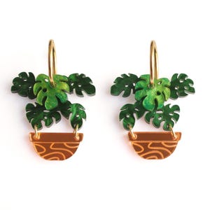 Può includere: Un paio di orecchini con un design a pianta di monstera verde. Gli orecchini hanno cerchi dorati e un vaso marrone con un motivo decorativo. Le foglie sono di un verde vibrante e il design generale è a tema botanico.