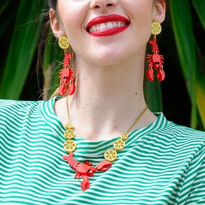 May include: A woman wearing a green and white striped shirt with a gold necklace and red lobster earrings with yellow lemon slices. The necklace features a lobster charm and lemon slices.