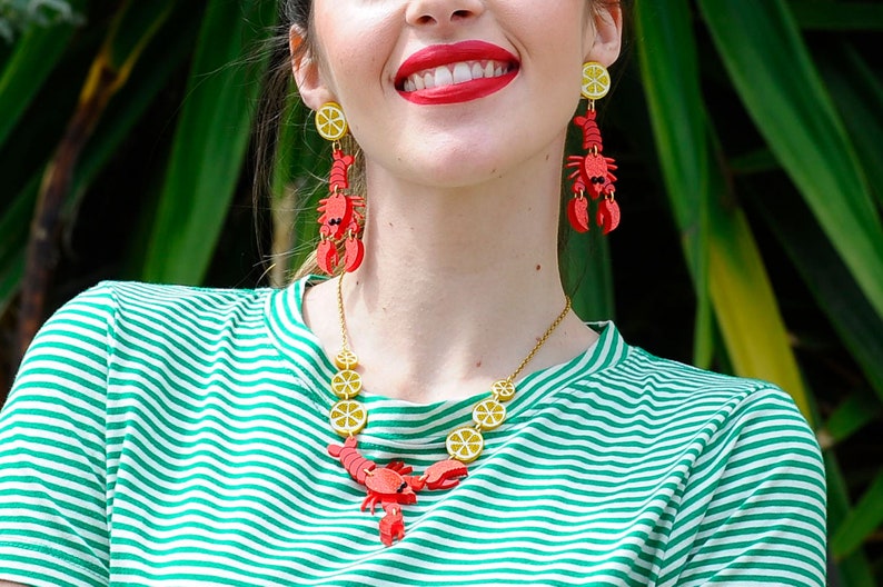 May include: A woman wearing a green and white striped shirt with a gold necklace and red lobster earrings with yellow lemon slices. The necklace features a lobster charm and lemon slices.