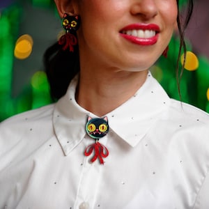 Peut inclure: Une chemise blanche à col avec de petits clous argentés est ornée d'une broche chat noire et de boucles d'oreilles assorties. Les accessoires pour chats ont des yeux jaunes et des nœuds rouges. L'arrière-plan est vert et jaune flou.