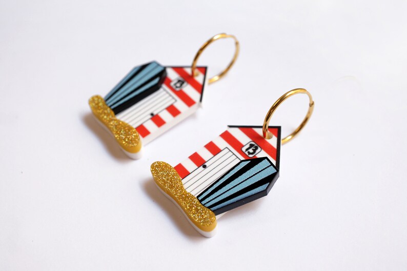 May include: A pair of gold hoop earrings with red and white striped beach hut charms. The huts have blue doors and windows and the number "13" is printed on the side.