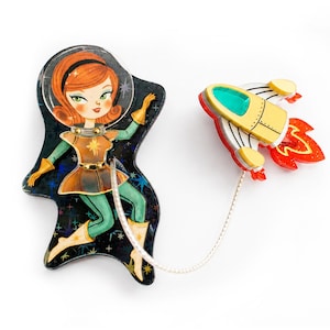 May include: A two-piece brooch set featuring a character with orange hair in a space suit on a black star-patterned background, connected by a silver chain to a yellow and white rocket ship with a red flame exhaust and a green window.