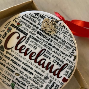May include: A white ceramic ornament with a red heart charm. The ornament features a word cloud of Cleveland landmarks, including the Rock and Roll Hall of Fame, the Browns, and the West Side Market. The ornament is a great way to show your Cleveland pride.