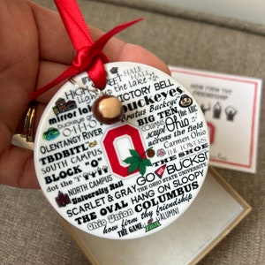 May include: White ceramic ornament with a red Ohio State University logo and the words "Go Bucks!" and "The Oval Columbus" in black lettering. The ornament is decorated with other Ohio State University-themed imagery, including the script "Ohio" and the words "Scarlet & Gray".