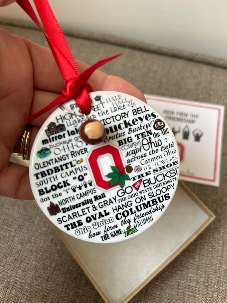 May include: White ceramic ornament with a red Ohio State University logo and the words "Go Bucks!" and "The Oval Columbus" in black lettering. The ornament is decorated with other Ohio State University-themed imagery, including the script "Ohio" and the words "Scarlet & Gray".